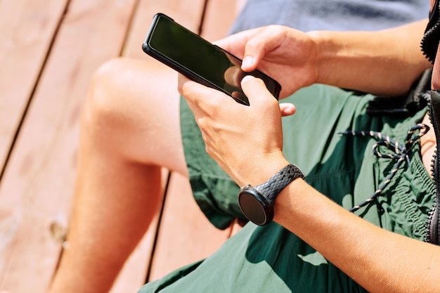 Hands of young sportsman in shorts scrolling in mobile phone while relaxing at break between outdoor sports trainings