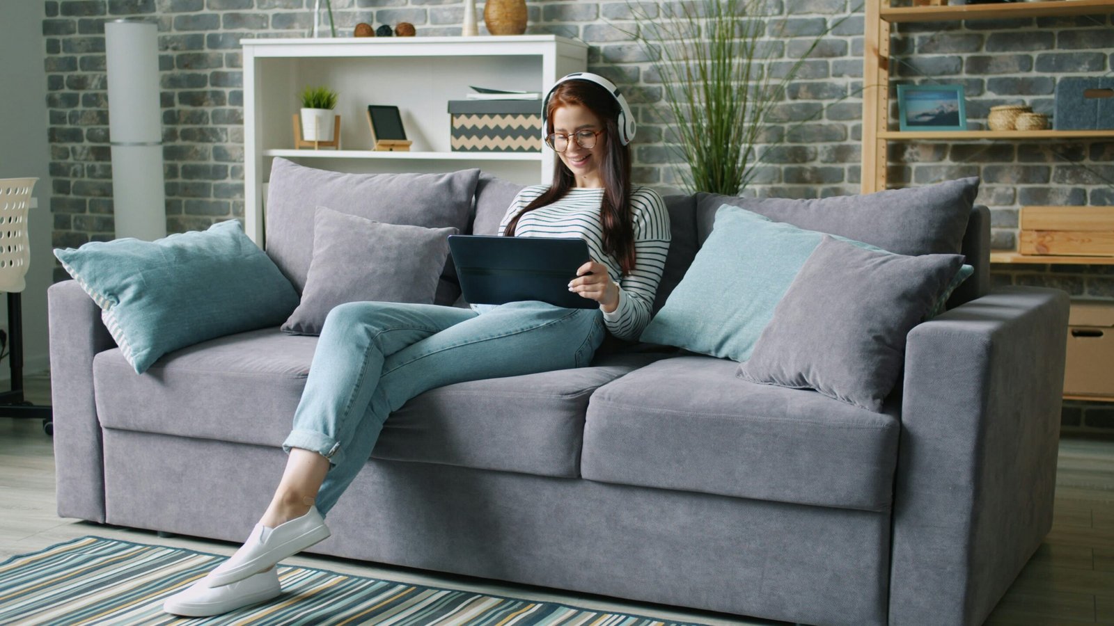 Woman wearing headphones uses tablet on couch.