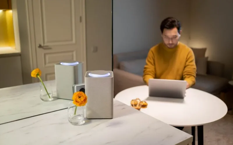Asian man at a table using a laptop with a smart speaker nearby, indoors.