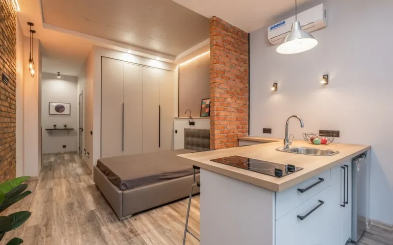 Miniature kitchen with modern appliances and bedroom behind brick partition in small loft style apartment