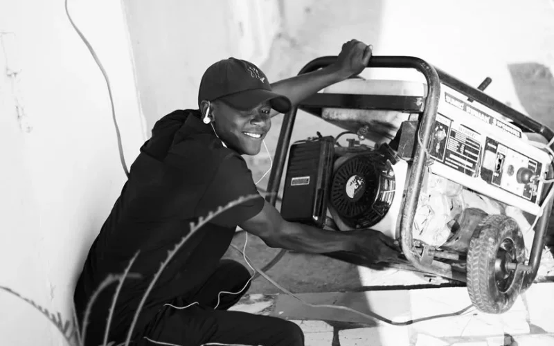 A cheerful young man wearing a cap and earphones operating a portable generator outside.