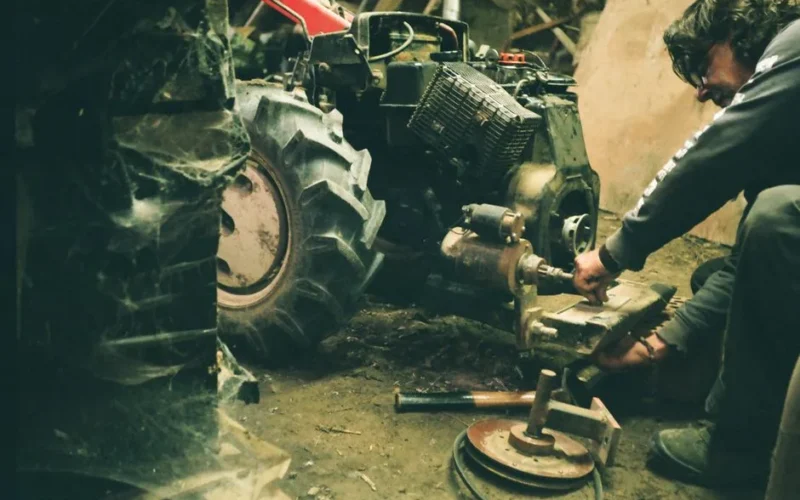 A mechanic working on a tractor's engine in a dimly lit indoor workshop.