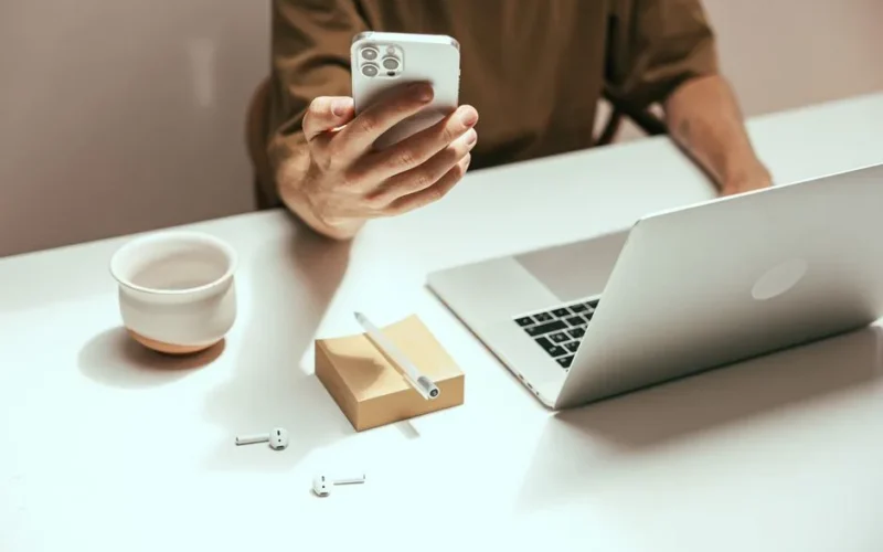 A person working remotely with a smartphone, laptop, and wireless earbuds on a desk.