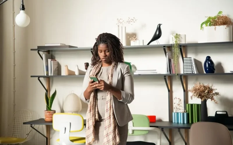 A woman standing in a modern office environment, focused on her smartphone, surrounded by books and plants.