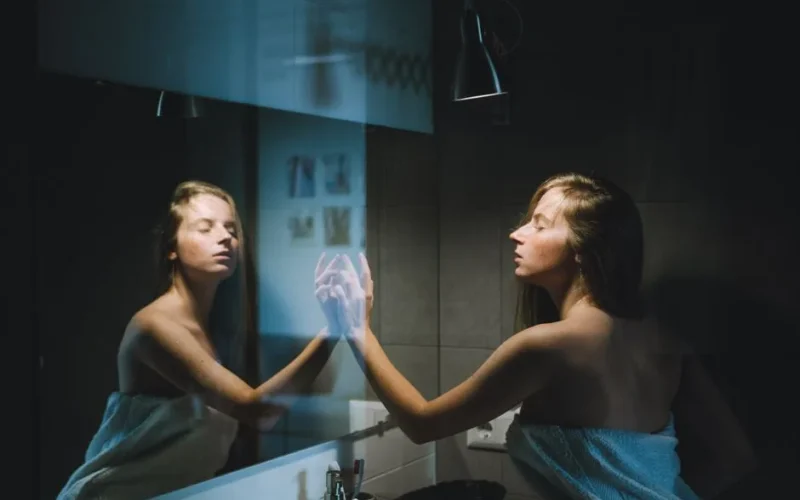 A young woman wrapped in a towel touches the mirror in a contemporary bathroom setting.