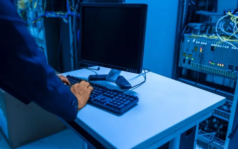 An IT professional operates a computer in a server room, managing network systems and connected devices.