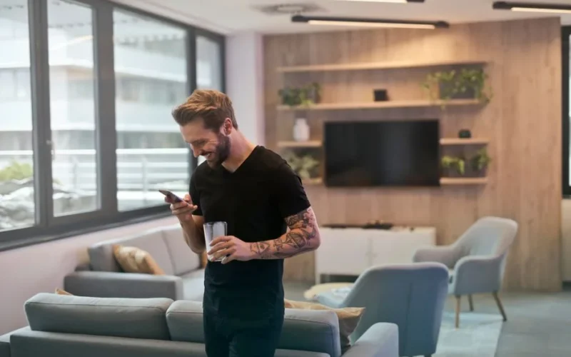 Casual man with tattoos laughing while texting on smartphone in modern office lounge.