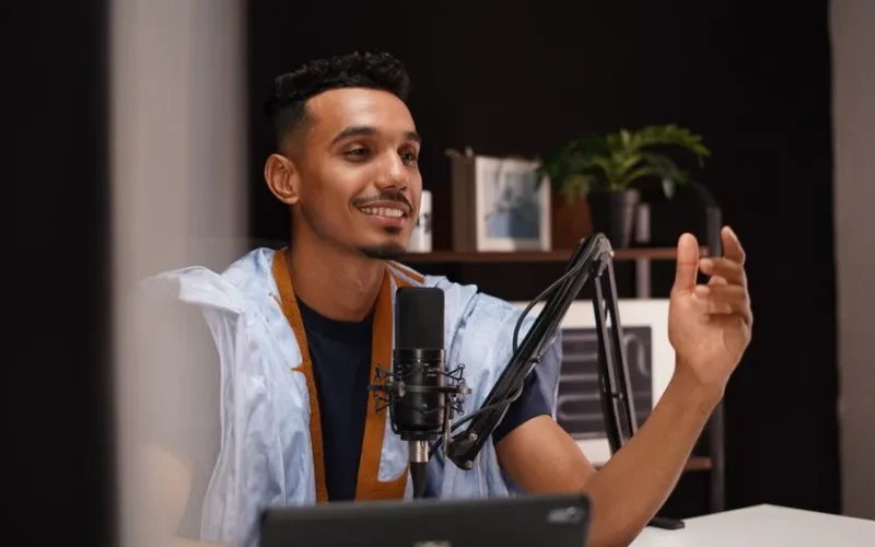 Smiling young man recording a podcast in a cozy home studio with professional microphone setup.