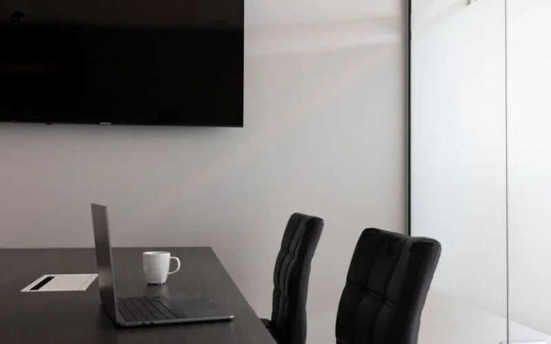 A sleek, modern office setup with a laptop, cup, and TV in an empty conference room.
