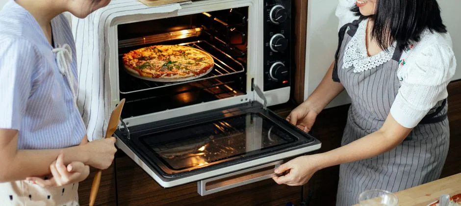 Side view of anonymous female cooks in aprons placing delicious homemade pizza in hot electrical oven while baking in kitchen