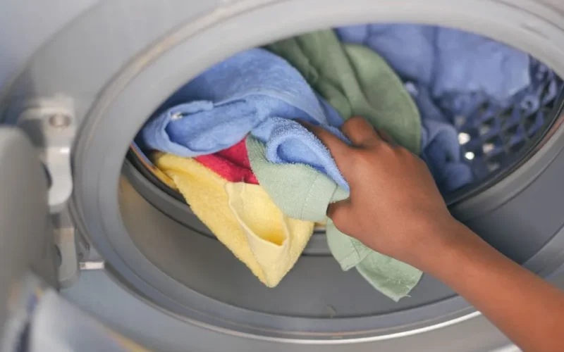 A close-up of a hand loading colorful towels into a front-loading washing machine.