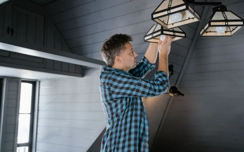 A man in a plaid shirt changes a lightbulb in a stylish attic room.