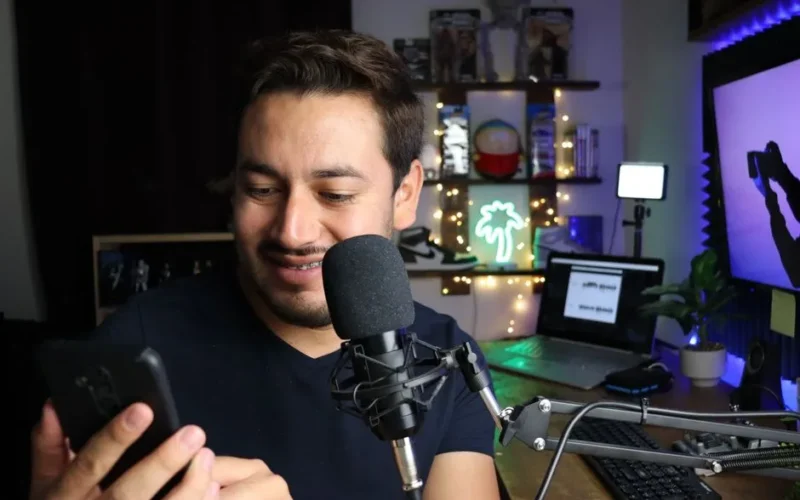 A man using a smartphone while seated at a desk with a microphone, surrounded by cozy decor.