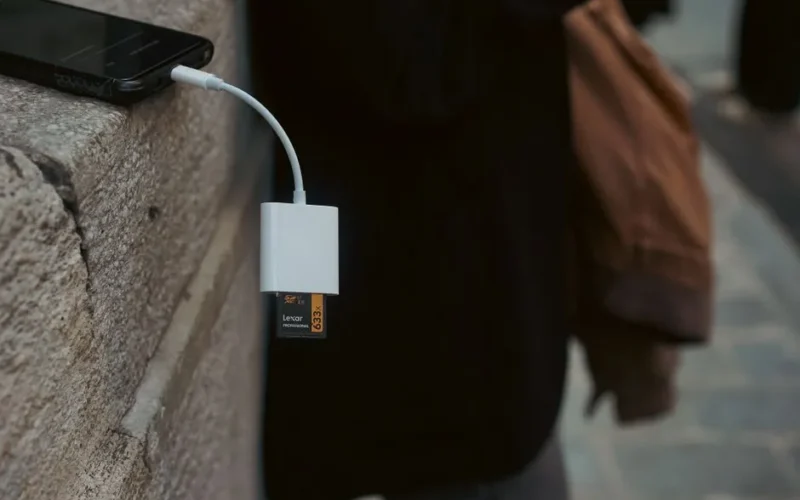 A mobile phone connected to a memory card reader outdoors on a stone surface.