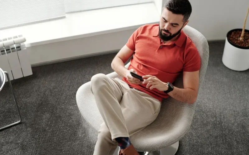 Man in red polo shirt using smartphone while sitting in a modern office chair.