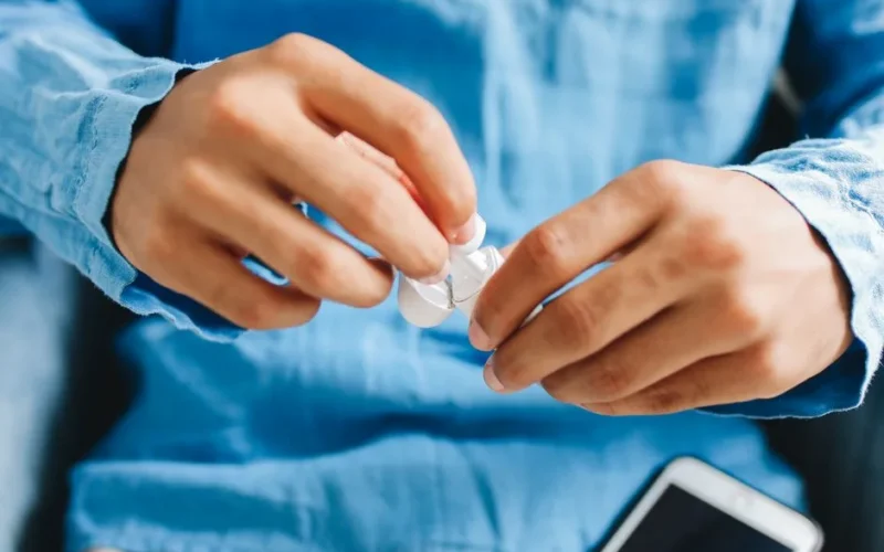 A man in a blue shirt holding wireless earbuds with a smartphone visible.
