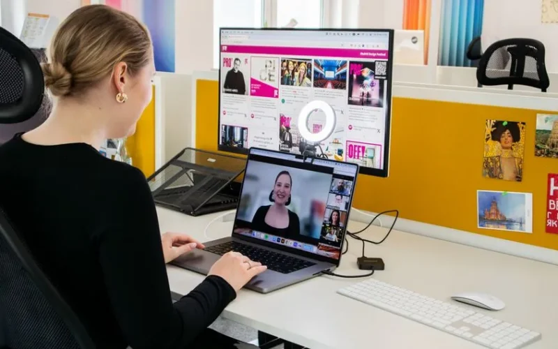 A woman conducts a professional video call while working at her desk in a bright office setting.