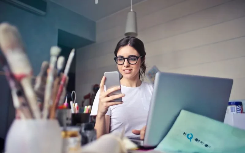 A woman using her phone at a desk, surrounded by art supplies and a laptop, in a creative workspace.