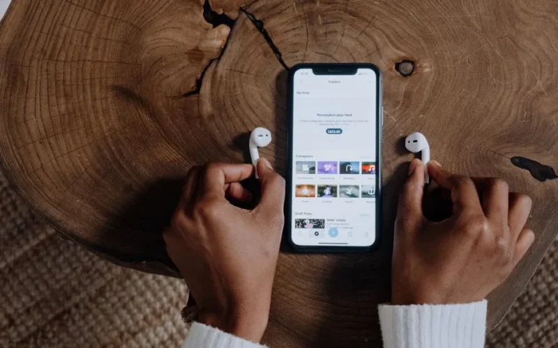 Hands holding wireless AirPods next to a smartphone displaying a social app on a wooden surface.