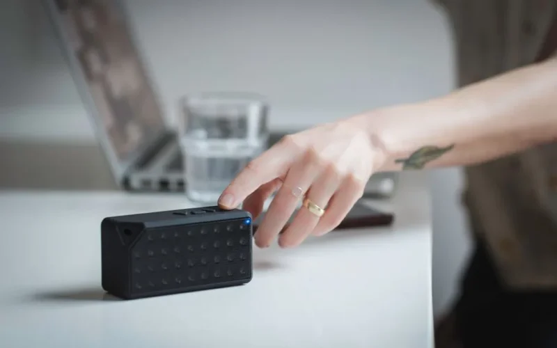 Close-up of a person adjusting a portable Bluetooth speaker on a desk indoors, with a laptop in the background.