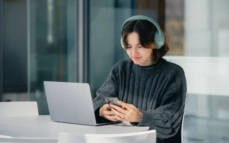A young woman wearing headphones uses a laptop and smartphone indoors at a bright table.