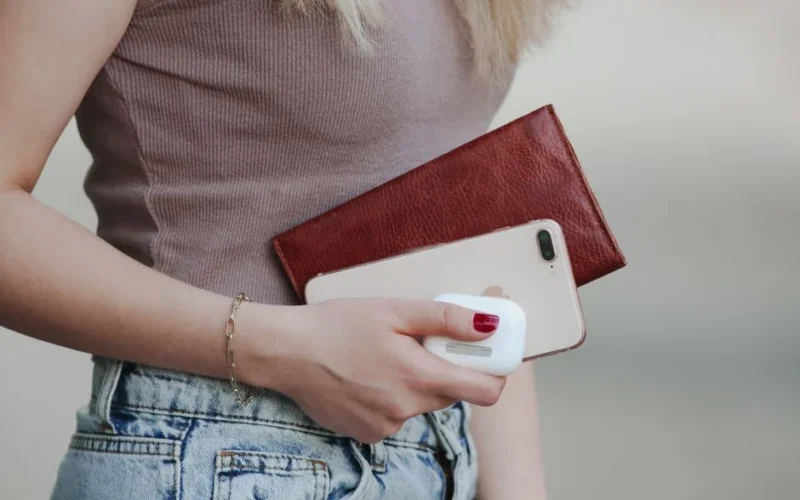 Woman holding smartphone, leather wallet, and wireless earbuds in casual attire.