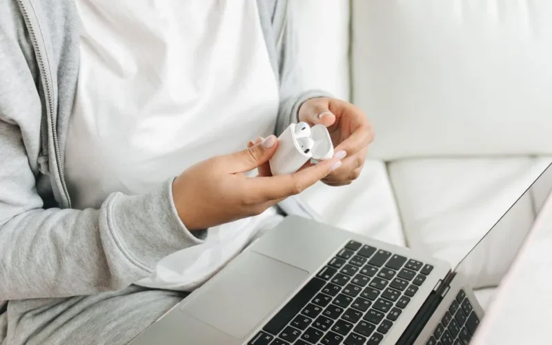 Close-up of hands opening wireless earbuds case near a laptop on a white couch.