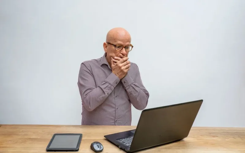Elderly man reacts in surprise to laptop screen at wooden desk with tablet and mouse.