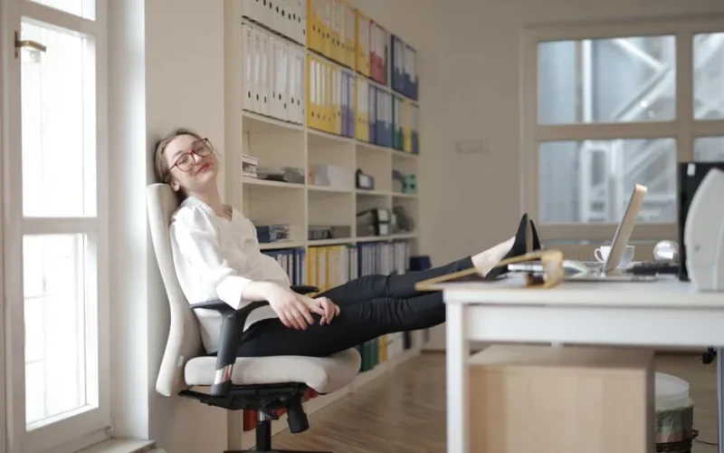 Side view of content female employee wearing formal clothes and eyeglasses sitting on chair with crossed feet on table and chilling during work with closed eyes in modern office