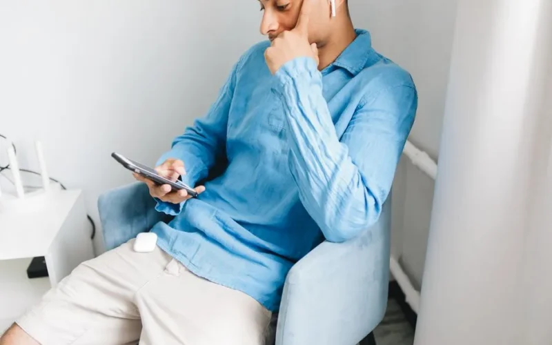 Young man in a blue shirt sits indoors, using a smartphone and wireless earbuds, enjoying music or a podcast.