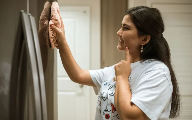 A woman cheerfully wipes a refrigerator, showcasing a moment of home cleaning.
