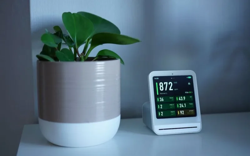 A modern air quality monitor next to a green plant on an indoor table, measuring CO2 levels.
