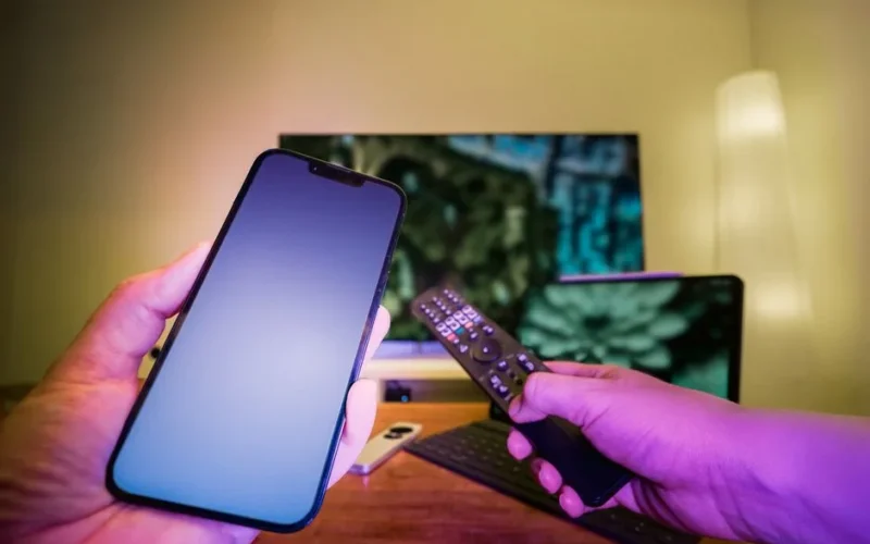 Hands holding a smartphone and remote control in a high-tech home setup with TV and other electronic devices.