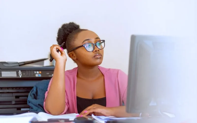 African woman in pink blouse working at her desk in an office with computer and printer.