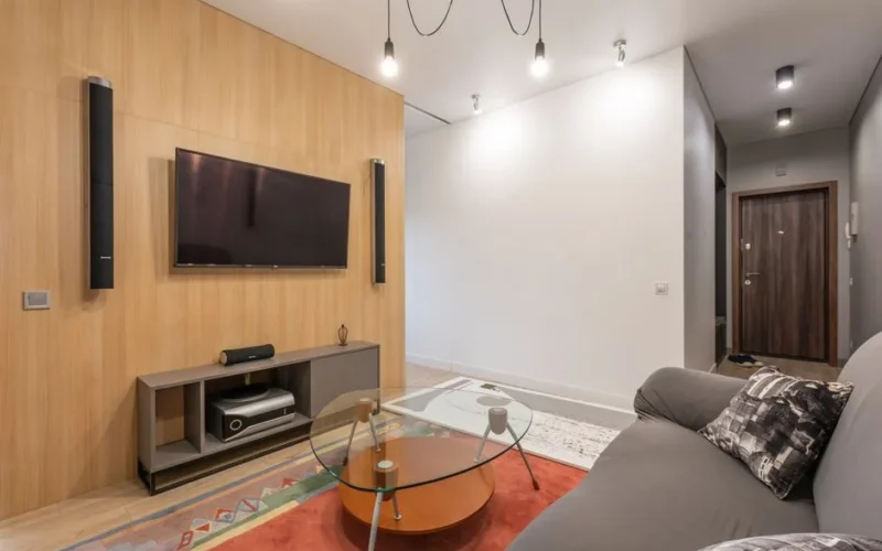Interior of contemporary light apartment with couch with pillows near table on carpet and cabinet with TV on wall with speakers near lamps on ceiling