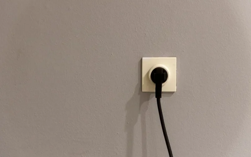 A black plug inserted into a white wall socket on a plain grey wall.