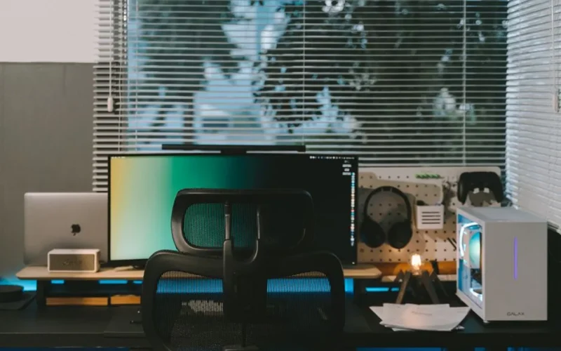 A stylish office desk setup featuring a computer, gadgets, and large windows with blinds.