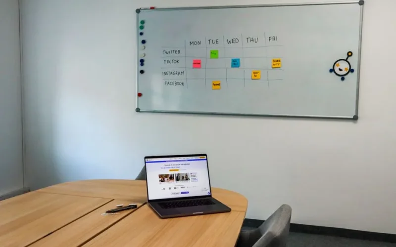 Office setup featuring a conference table, laptop, and organized whiteboard schedule, highlighting a tech-driven workspace.