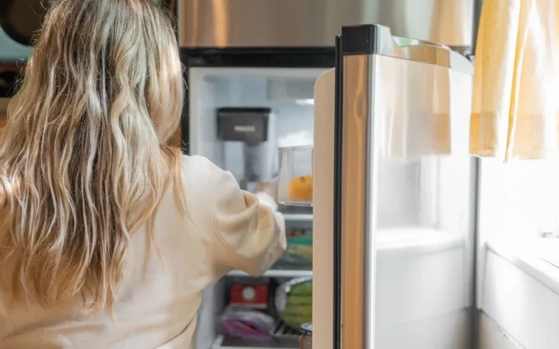Back view of a blonde woman retrieving food from a refrigerator in a bright kitchen.