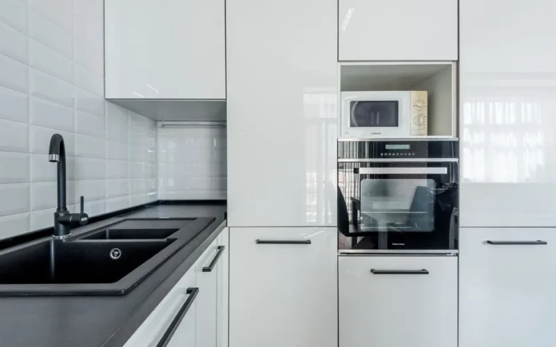 Interior of light black and white kitchen corner with white cupboards and black sink near white tiled wall and oven with microwave