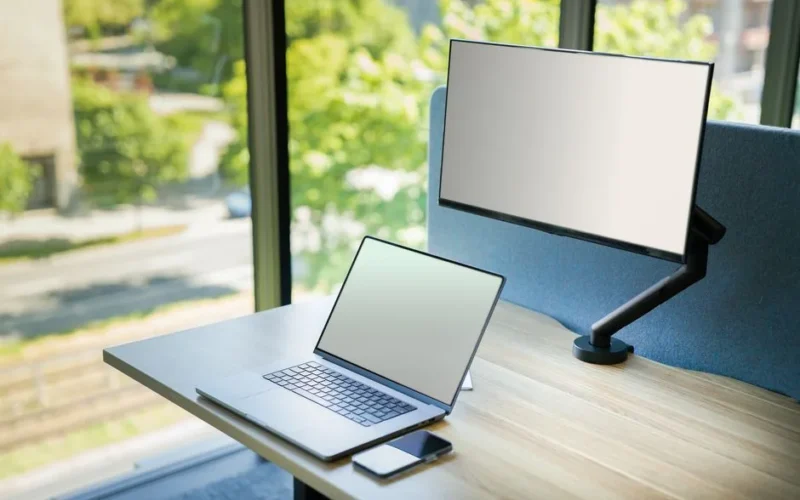 A contemporary office desk setup with a laptop and monitor by a window, featuring bright natural light.