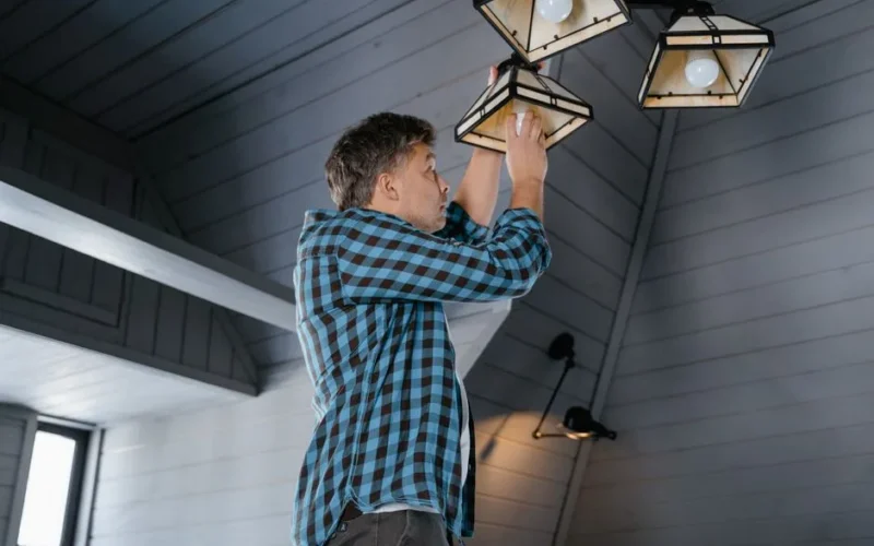 A man in a plaid shirt changes a light bulb on a ceiling lamp in a stylish, modern interior.