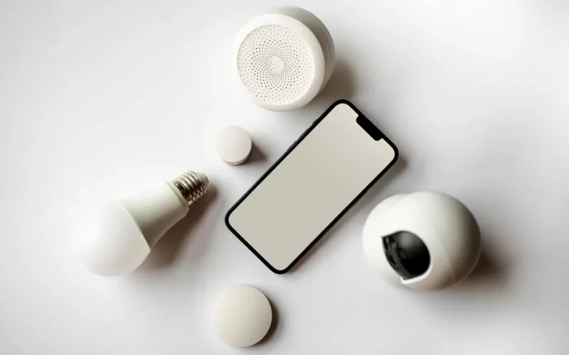 Flat lay of smartphone and smart home devices on a white backdrop, showcasing modern technology.