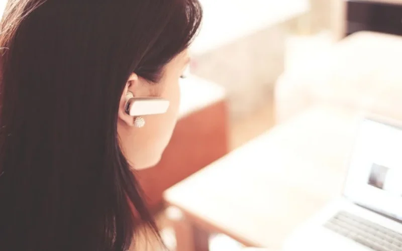 A businesswoman using a Bluetooth headset while working on her laptop in a modern office.