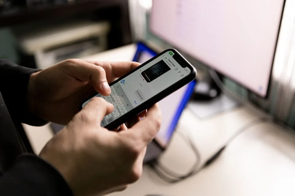 Osoba trzymająca smartfon i pisząca wiadomości w pomieszczeniu przy ekranie komputera